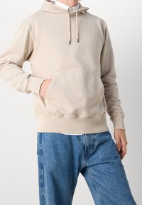 Beige hoodie med dragsko vid huvan, känguruficka, ribbade muddar och fåll; bärs med blå denimjeans och visar en avslappnad passform.