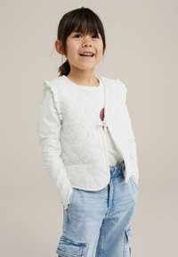 Vit quiltad väst med volangdetaljer på axlarna, bärs över en långärmad vit skjorta. Blå denim cargobyxor med fickor.