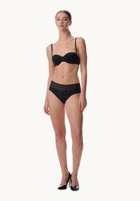 Zwart gewatteerd balconette-bh met dunne bandjes en zwarte slip met hoge taille en een inzetstuk van gaas. Model staat op zwarte hakken voor een lichte achtergrond.