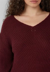 Bordeaux gebreide trui met V-hals en een gestructureerd patroon, voorzien van geribde details en subtiele stiksels langs de halslijn.