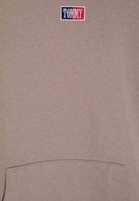 Felpa marrone chiaro con tasca a marsupio, dotata di un piccolo logo ricamato "TOMMY" in blu e rosso nella parte alta a sinistra. Tessuto morbido e texturizzato.