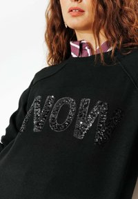 Sweatshirt noir avec un grand texte "NOW" en sequins brillants, col rond et un col côtelé avec une chemise rayée visible en dessous.