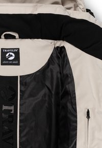 Beige en zwarte reisk jacket met zwarte binnenvoering, een ritszak en een label "Travelin' where we walk" in het kraaggebied.