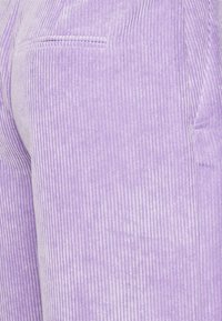 Lila kordfløyelsbukser med vertikale striper, med en subtil glans og detalj med sidelommer. Stoffet ser teksturert og mykt ut.