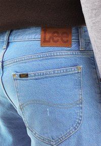 Jeans in denim blu chiaro con un'etichetta in pelle marrone "Lee" sulla cintura. Presentano tasche posteriori classiche con cuciture gialle.