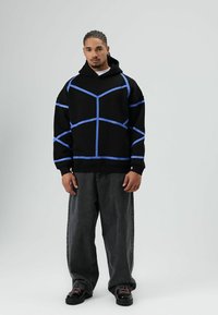 Zwarte oversized hoodie met blauwe geometrische strepen, lange mouwen, geribbelde manchetten, gecombineerd met losse zwarte jeans en zwarte schoenen.