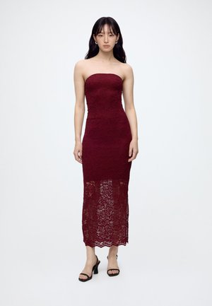 Femme portant une robe bustier en dentelle bordeaux avec une jupe inférieure transparente, debout devant un fond blanc uni.