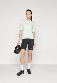Licht mintgroene korte mouwen fietstop gecombineerd met zwarte padded shorts. Het model houdt een zwarte helm vast en draagt witte sokken en zwarte fietschoenen.