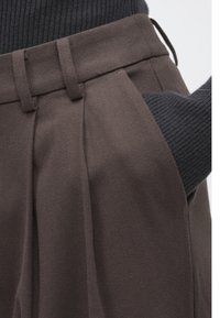 Pantalones de tela marrón con pliegues laterales; incluye un detalle de bolsillo. La pretina es lisa con trabillas para el cinturón. Se ve un suéter texturizado de color gris oscuro.