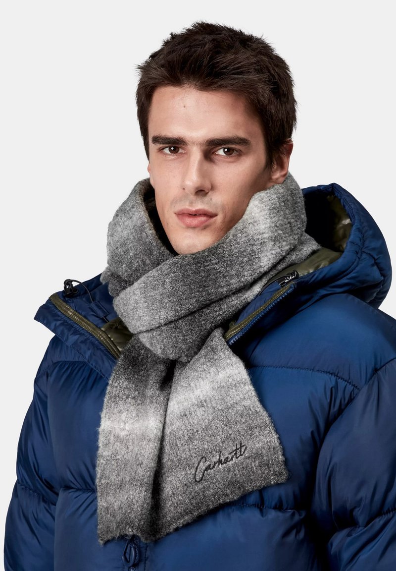 Bufanda de lana gris con sombreado degradado, que presenta una textura suave y un logo bordado. Se lleva sobre una chaqueta acolchada azul con capucha.