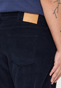 Navy corduroy trousers with a textured finish, featuring a leather label at the waistband and a back pocket with a subtle curve.