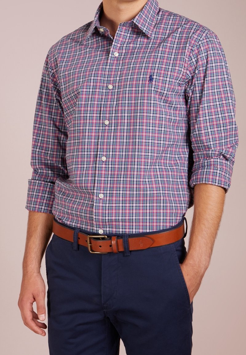 Rutigt skjorta i röd, blå och vit, med långärmar som är uppkavlade. Kombinerad med mörknavy byxor och ett brunt läderbälte.