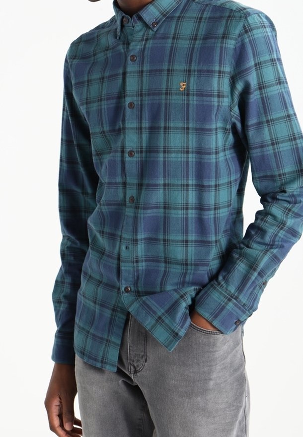 Man in een teal en marineblauwe geruite overhemd met geborduurd logo, in combinatie met lichtgrijze jeans, met hand in de zak.