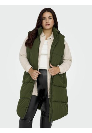 Vihreä pehmeä liivi, jossa on korkea kaulus ja sivutaskut, päällä avoimena vaaleanbeigen nappipaidan ja mustien legginsien kanssa, jonka pinta on sileä.