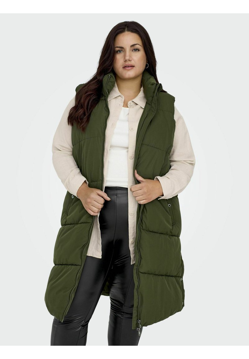 Chaleco acolchado verde con cuello alto y bolsillos laterales, llevado abierto sobre una camisa de botones beige claro y leggings negros, con una textura suave.