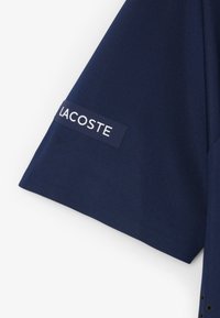 Tessuto blu navy con una toppa rettangolare con la scritta "LACOSTE" in lettere bianche sulla manica.