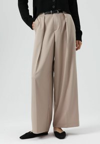 Beige wijde broek met voorplooien, gemaakt van gladde stof. Gestyled met een zwarte riem en gecombineerd met zwarte instapschoenen.