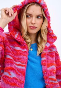 Futrzana kurtka z kapturem w kolorze różowym, czerwonym i lawendowym z falistymi wzorami; wyposażona w zamek błyskawiczny i miękką teksturę. Noszona na niebieskiej bluzce.