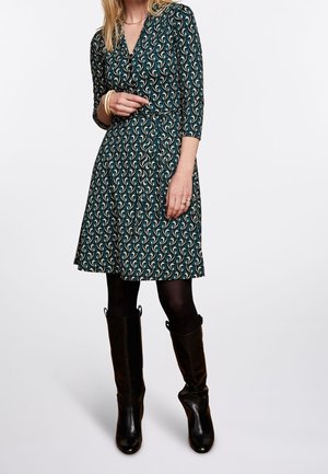 Femme portant une robe mi-longue à motifs vert foncé avec des manches trois-quarts, des collants noirs et des bottes noires montantes jusqu'aux genoux.