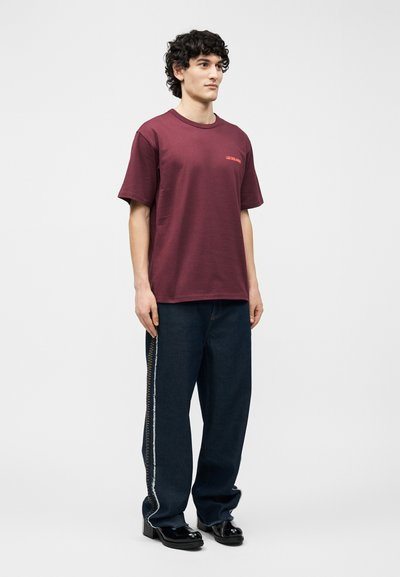 Νεαρός άνδρας με σγουρά μαλλιά, που φοράει μια φαρδιά μπορντό μπλούζα με μικρό κείμενο, σκούρο τζιν με φαρδιές μπατζάκια και πλαϊνή ραφή, και μαύρα γυαλιστερά παπούτσια.