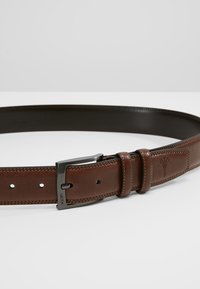 Ceinture en cuir brun avec une texture lisse, une boucle en métal argenté et un logo en relief subtil. Elle présente des coutures et des trous ajustables.