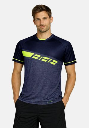 Mann steht mit den Händen in den Taschen, trägt ein sportliches Shirt in Marineblau und fluoreszierendem Gelb sowie schwarze Shorts vor neutralem Hintergrund.