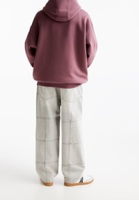 Persona di spalle che indossa un ampio hoodie bordeaux, pantaloni grigi chiari a gamba larga con cuciture e scarpe bianche con suola color gomma su sfondo bianco.