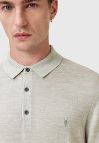 Lys grå polo t-shirt med en struktureret finish, tre sorte knapper og et broderet logo på brystet. Blød krave med en afslappet pasform.