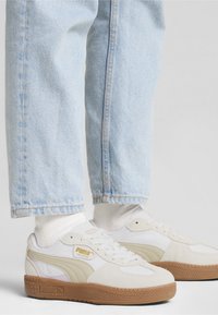 Baskets en daim blanc et beige avec une semelle en gomme, présentant une finition texturée et un logo doré en accent, associées à un jean bleu clair.