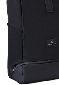 Primo piano di uno zaino Johnny Urban nero che mostra la tasca laterale, il compartimento inferiore con cerniera e la toppa con logo sul pannello frontale.