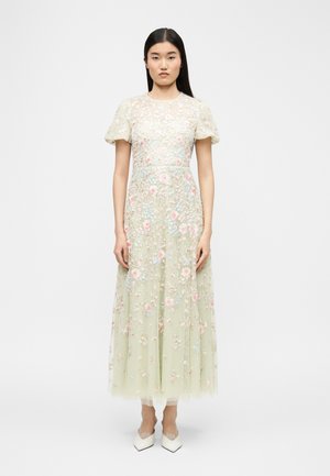 Femme debout, vêtue d'une robe midi vert clair brodée de fleurs, avec des manches courtes bouffantes et des chaussures blanches à bouts pointus, sur un fond uni.