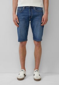 Shorts en denim bleu foncé avec un design classique à cinq poches, longueur genou, texture délavée et ourlets cousus, associés à des baskets blanches.