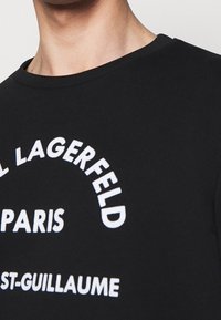 Persona che indossa una maglietta nera con testo curvo bianco che recita "KARL LAGERFELD PARIS ST-GUILLAUME" sul petto.