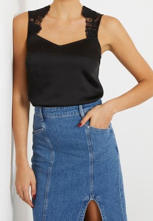 Vrouw draagt een zwarte mouwloze top met kanten bandjes, ingestopt in een hooggetailleerde blauwe denimrok met vooropenening en zakken.