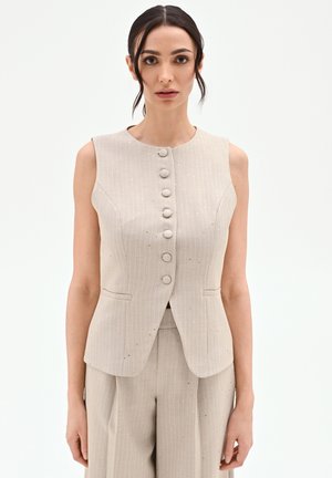 Donna che indossa un gilet smanicato beige a righe con bottoni, abbinato a pantaloni larghi coordinati, in piedi davanti a uno sfondo semplice.