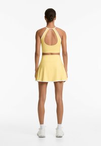 Top sportiv galben cu un design cu spatele deschis și o fustă evazată asortată, având un tiv alb. Textură netedă, material elastic.