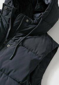 Černá prošívaná vesta s vysokým límcem, vpředu se zipem, kapucí na stažení a druky. Hladká, lesklá tkanina s prošívanými částmi.