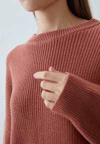 Gestrickter Pullover in Rosé mit weitem Ausschnitt, gerippten Bündchen und strukturiertem Stoff, zeigt Handgeste nahe dem Stoff.