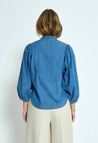 Džínová košile s nafouklými rukávy, zakřiveným lemem a klasickým límečkem. Materiál má jemnou texturu v středně modrém odstínu.