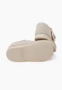 Zapatos de lona beige sin cordones con una suela de goma plana y un detalle de botón lateral. La parte superior texturizada tiene un aspecto tejido.