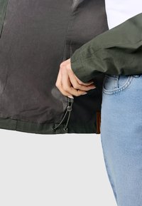 Personne ajustant le cordon de serrage à l’ourlet inférieur d’une veste vert foncé tout en portant un jean bleu clair.