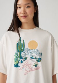 Camiseta blanca de algodón con un gráfico colorido de paisaje desértico que incluye cactus, montañas y un sol, acompañado del texto "Levi Strauss" en color rosa.