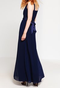 Kvinna med röd hår som bär en ärmlös marinblå golvlång klänning med en satinrosett knuten vid midjan, stående i svarta högklackade skor.