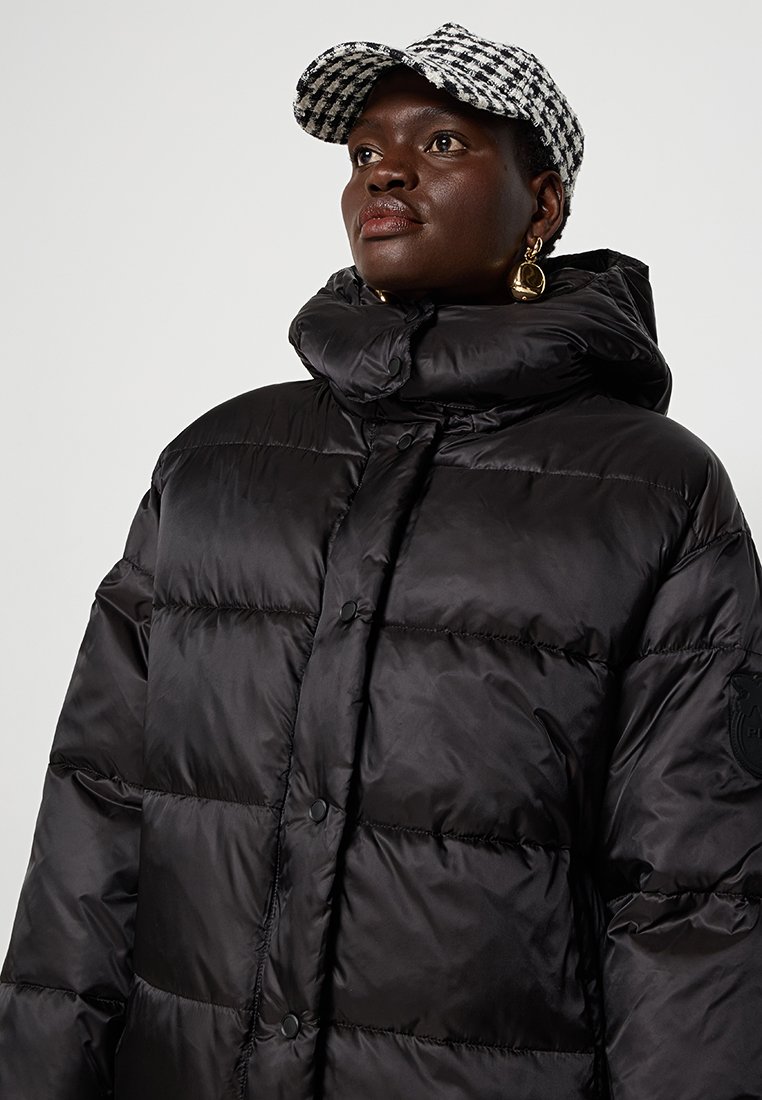 Schwarze Daunenjacke mit hohem Kragen, Druckknöpfen und einem Abzeichen-Detail am Ärmel. Getragen mit einer schwarz-weißen karierten Mütze.
