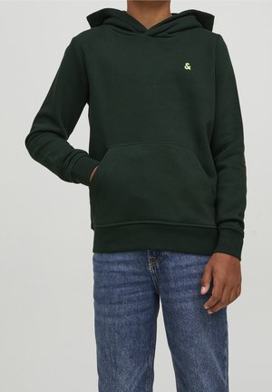 Person iført en mørkegrøn hættetrøje med en forlomme og et lille gult logo, kombineret med blå jeans, stående med den ene hånd i lommen.