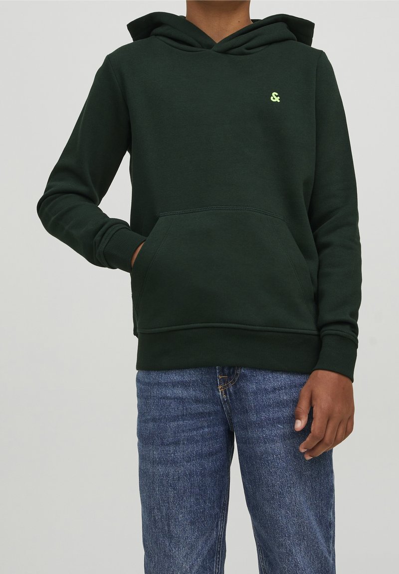 Personne portant un hoodie vert foncé avec une poche avant et un petit logo jaune, associé à un jean bleu, debout avec une main dans la poche.