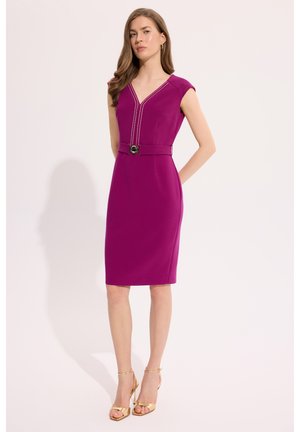 Kobieta stoi z rękami za plecami, ubrana w dopasowaną fioletową sukienkę do kolan z dekoltem w serek i paskiem, w zestawieniu ze złotymi sandałami na wysokim obcasie.