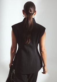 Zwart op maat gemaakte vest met gestructureerde schouders, aansluitende silhouet en subtiele naaddetails; gedragen over een broek en aangevuld met een leren tas.