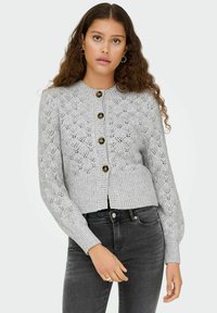 Svetlo siva pletena jopica z teksturo, pet temnih gumbov, rebrastimi manšetami in kratko zasnovo, v kombinaciji z črnimi jeans hlačami.