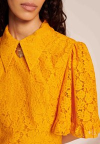 Blusa de renda amarela com colarinho clássico, padrão floral intricado, mangas bufantes e um broche decorativo em forma de coração na gola.